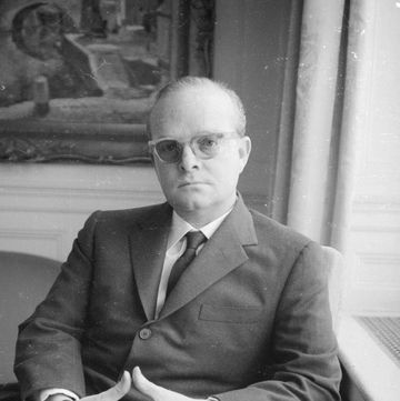 truman capote sits in an armchair and looks at the camera, he wears a suit with a tie, glasses, and a watch, his hands are clasped on his lap