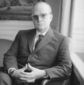 truman capote sits in an armchair and looks at the camera, he wears a suit with a tie, glasses, and a watch, his hands are clasped on his lap