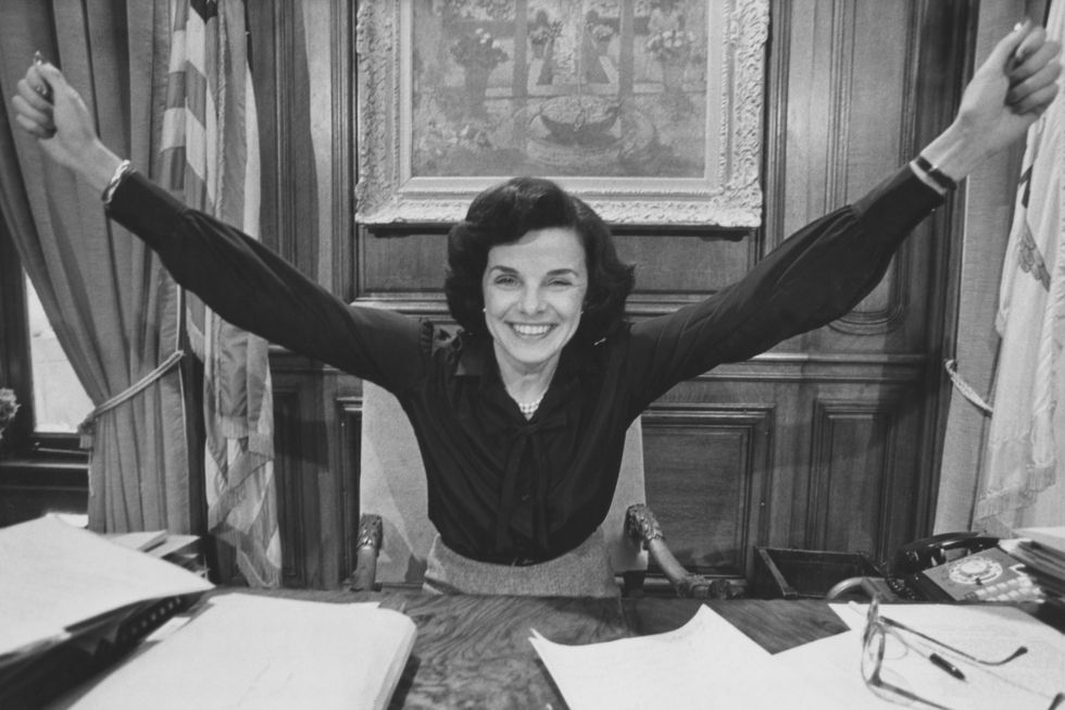 a black and white photo of dianne feinstein stretching her arms in victory and smiling as she sits at a desk in an office