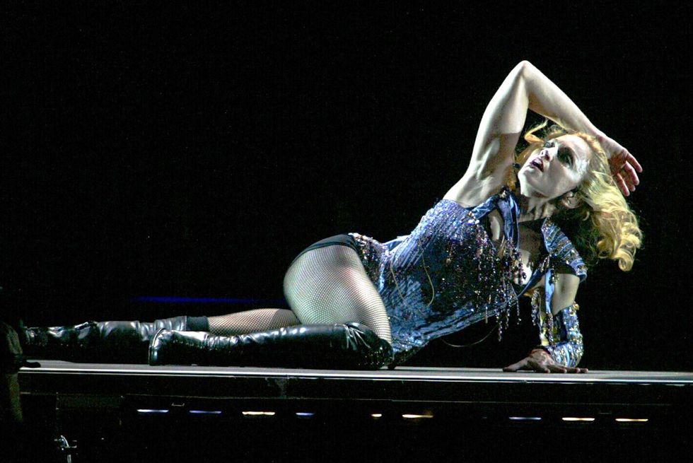 madonna, wearing a blue outfit, fishnet stockings, and black leather boots, lies on a stage while looking up, with her right arm resting on her head