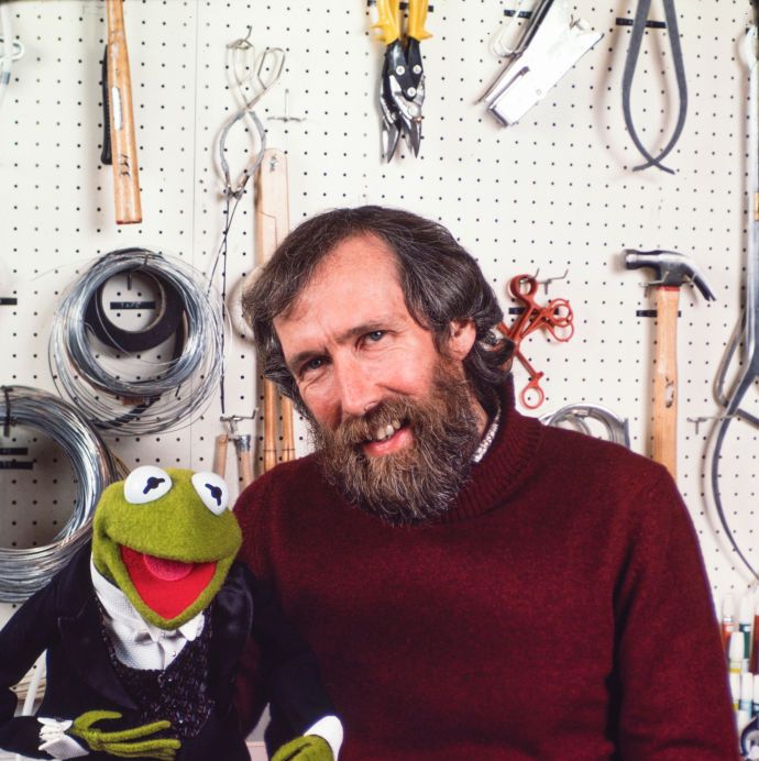 jim henson smiles at the camera as he holds kermit the frog on one hand, he stands in a workshop and wears a red sweater