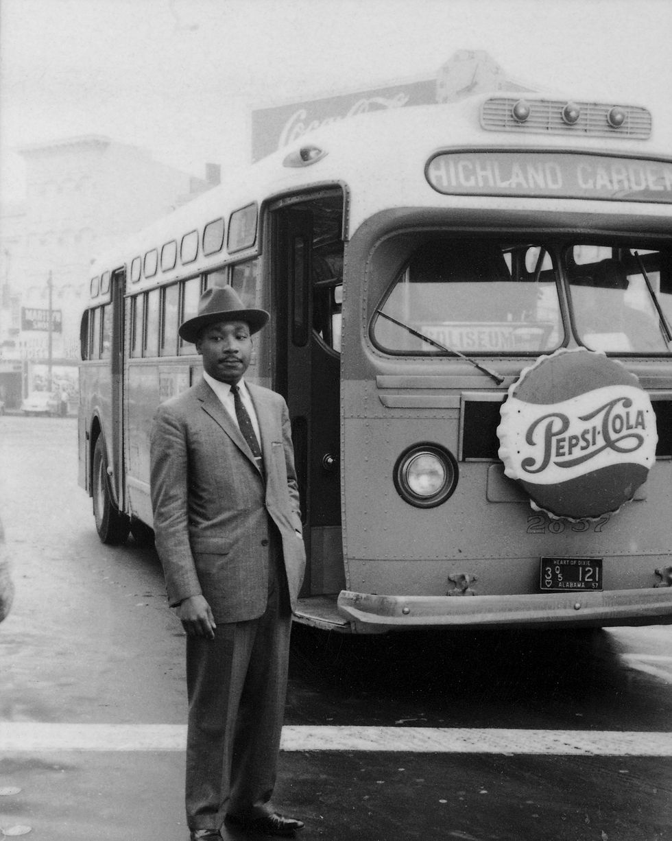 martin luther king jr stands outside in a suit and hat, behind him is a city bus with a pepsi cola ad on the front