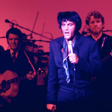 a man stands with his eyes closed and sings into a microphone he holds in one hand, two musicians play behind him