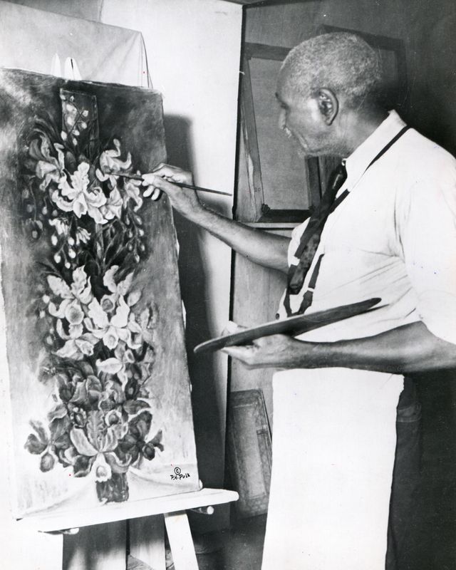 George Washington Carver pinta un cuadro floral sobre un lienzo mientras sostiene una paleta en una mano y un pincel en la otra.