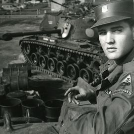 a man looks over his shoulder at the camera, he wears a military uniform and hat, behind him is a tank