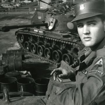 a man looks over his shoulder at the camera, he wears a military uniform and hat, behind him is a tank