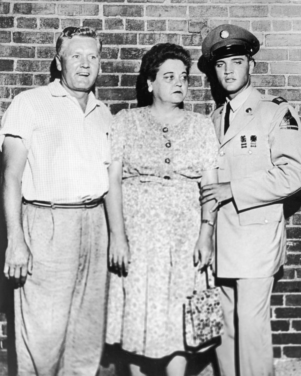 a woman stands between two men in front of a brick wall, the man on the right wears a military uniform and hat