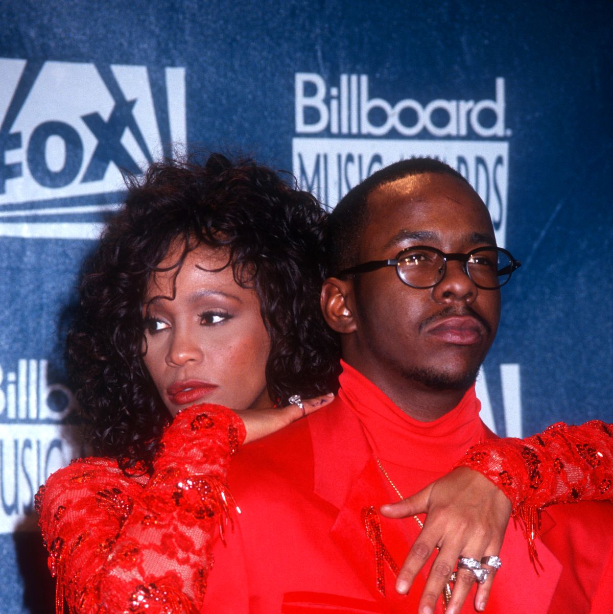 whitney houston and bobby brown pose for a photo, she hugs him from behind with her arms on his shoulders and chest, they wear matching bright red outfits