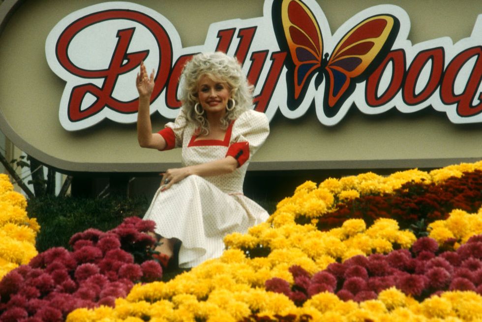 dolly parton wearing a white and red dress, sitting down in front of a large dollywood sign and waving, with several red and yellow flowers in front of her