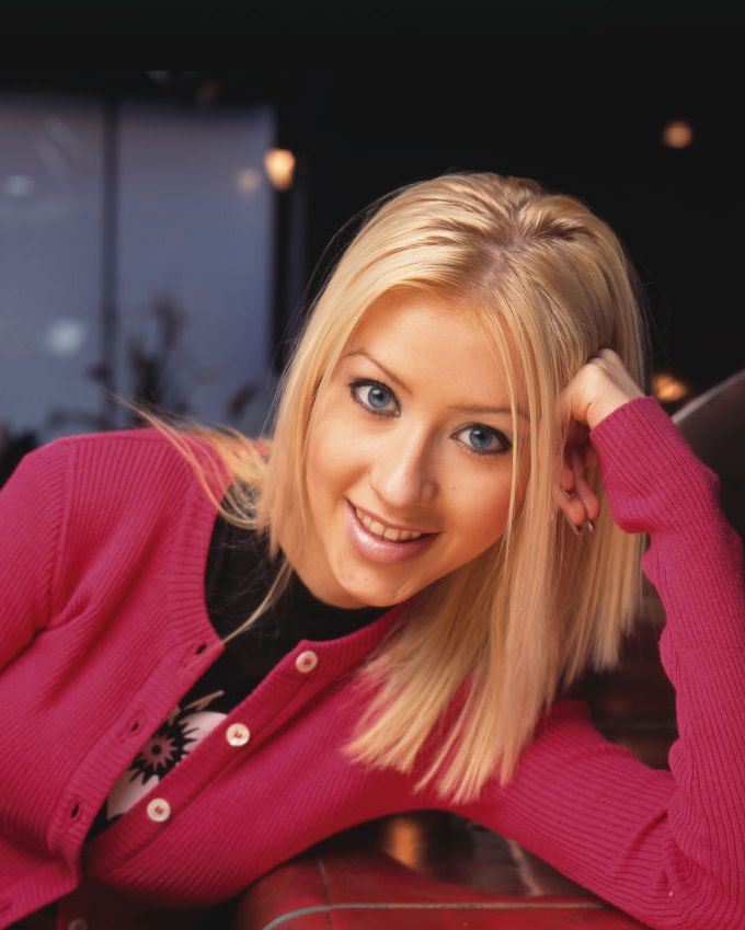 christina aguilera smiles at the camera while leaning her head against on hand, she wears a pink cardigan over a black shirt with a floral design on it