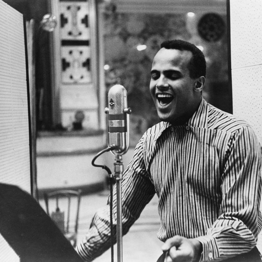 harry belafonte sings into a microphone inside a recording studio, he is wearing a striped collared shirt and his eyes are closed