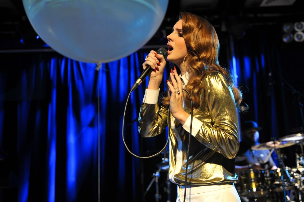 lana del ray sings into a microphone she holds in one hand, she stands on a stage and has her other hand in front of her, she wears a gold shimmering shirt with white cuffs and collar