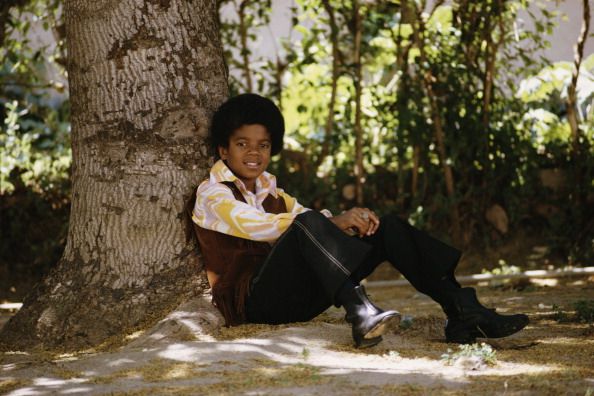 a young michael jackson wearing a vest, long sleeve shirt, black pants, and boots, sitting on the ground outside against a tree