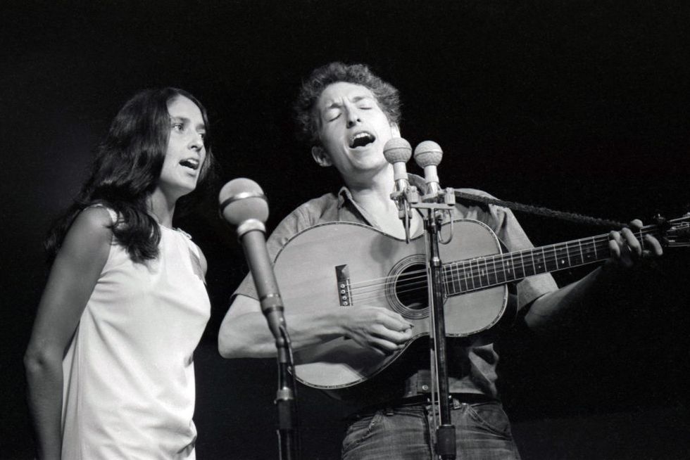 joan baez and bob dylan stand next to each other and sing into microphones on stands, he also plays guitar