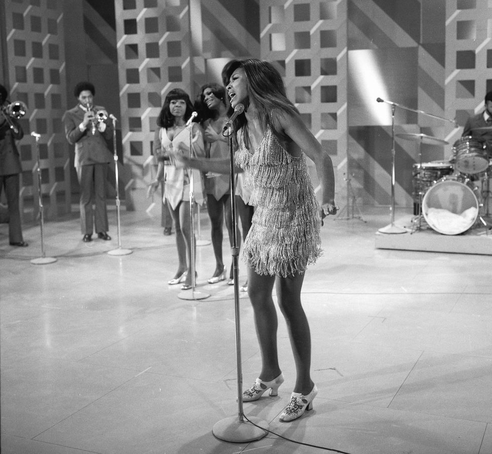tina turner sings into a microphone on a stand as backup singers an musicians perform behind her