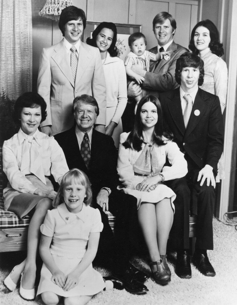 rosalynn and jimmy carter sit with their four children, amy who is sitting on the floor, donnel jeffrey aka jeff who is sitting to the right of the couple, james earl carter iii aka chip who is standing and john william aka jack who is also standing, the carters three daughter in laws are also in the photo, annette, caron and judy