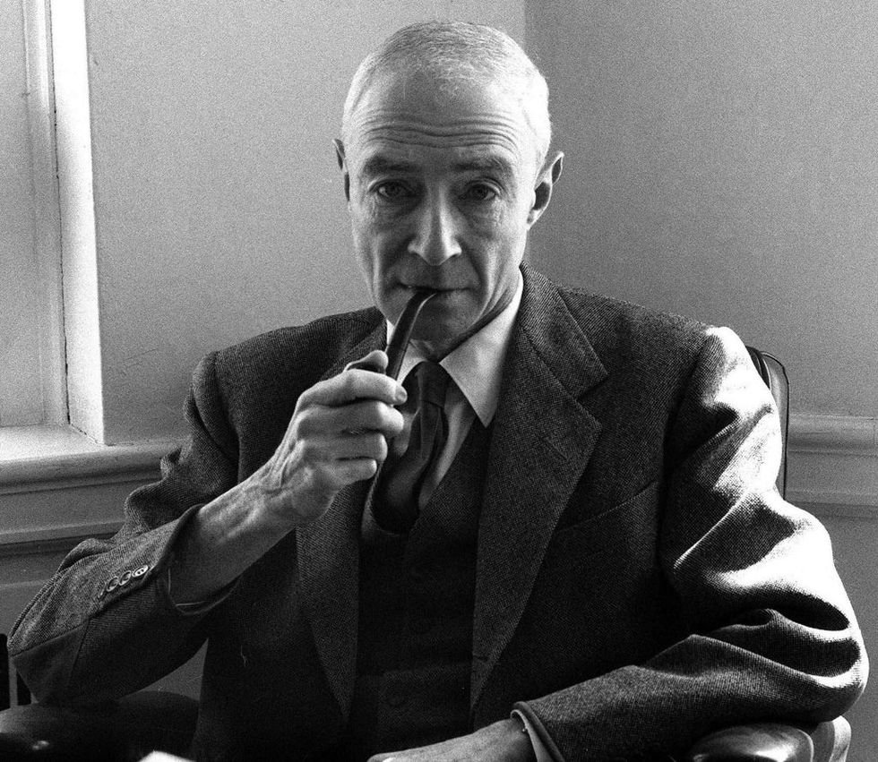 j robert oppenheimer wearings a suit and tie, and holds a pipe in his mouth