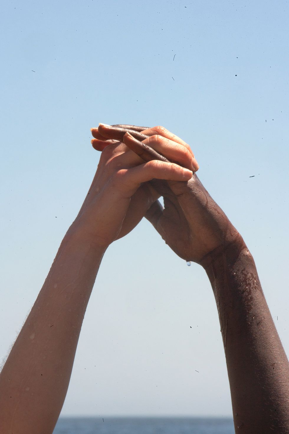 intermeshed fingers of two hands of separate people, with droplets of water dripping off the one, close to the wrist, in a show of togetherness, mutual affection solidarity with a rich blue sky for background