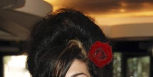 amy winehouse smiles at the camera, she wears a black strapless top with large white hoop earrings and a red rose in her beehive hairdo