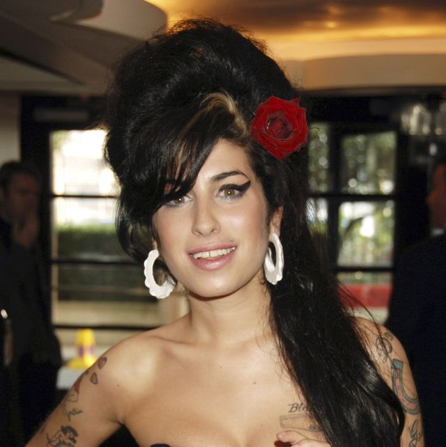 amy winehouse smiles at the camera, she wears a black strapless top with large white hoop earrings and a red rose in her beehive hairdo