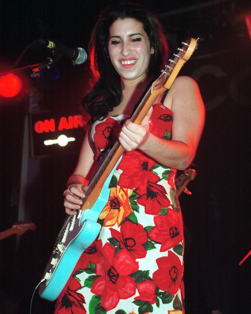 amy winehouse smiles while standing behind a microphone and playing guitar, she wears a floral pattern sleeveless dress
