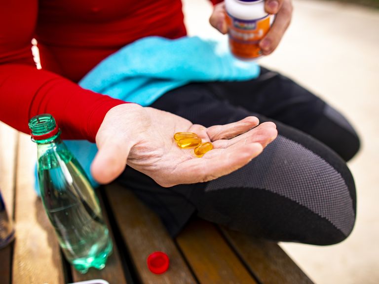 an athletic man holding out a couple of orange colored capsules on the palm of his hand while sitting on a park bench