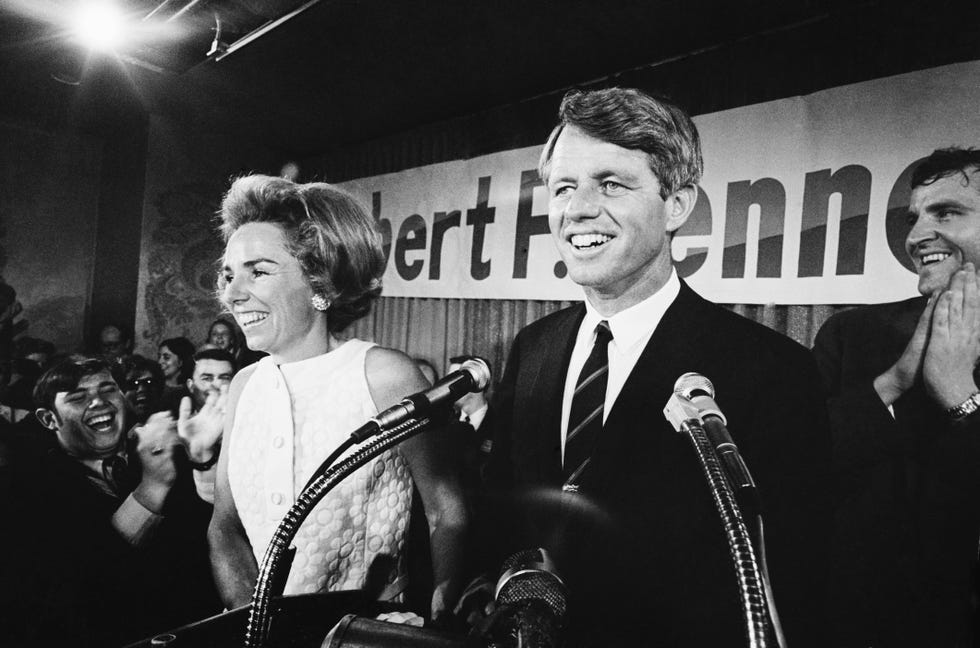 ethel kennedy and robert kennedy smile while standing near a podium with microphones, people smile and clap in the background