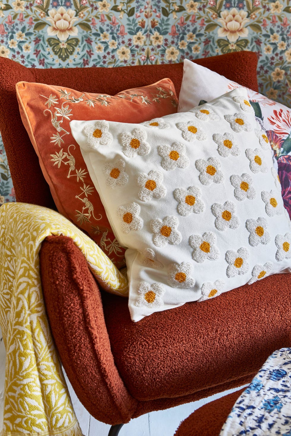 an orange boucle armchair with three patterned cushions and a yellow throw blanket on it, and blue floral wallpaper behind it