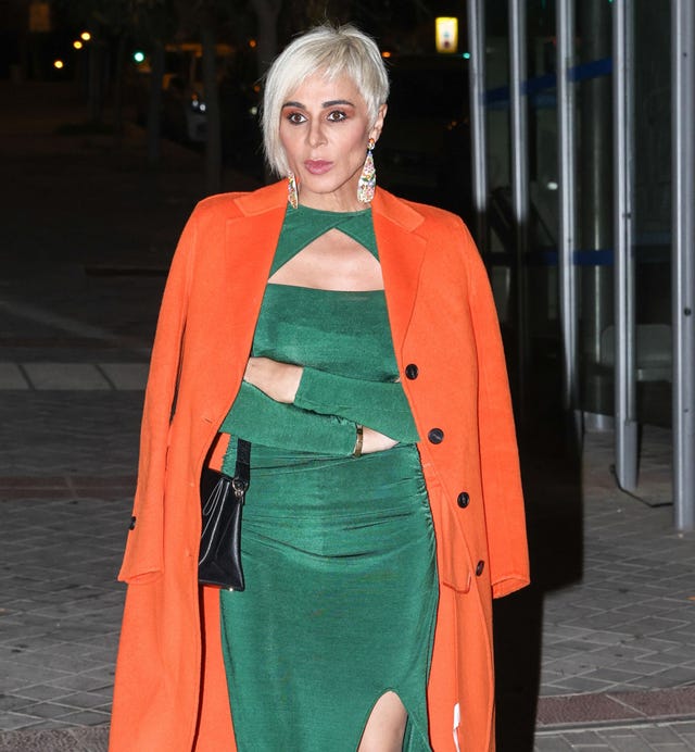 la diseñadora con abrigo naranja y vestido verde camina con los brazos cruzados