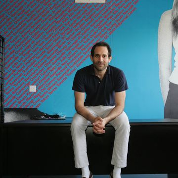 man sitting on a black table in an office with his hands folded, he stares at the camera and smiles