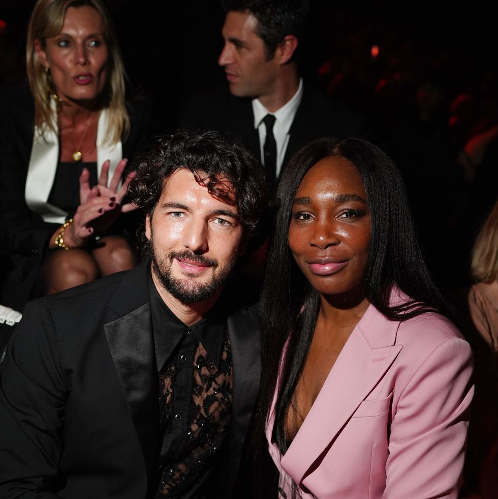 a man and a woman sit closely together and smile for a photo, he wears a black suit coat and transparent lace shirt, she wears a pink suit