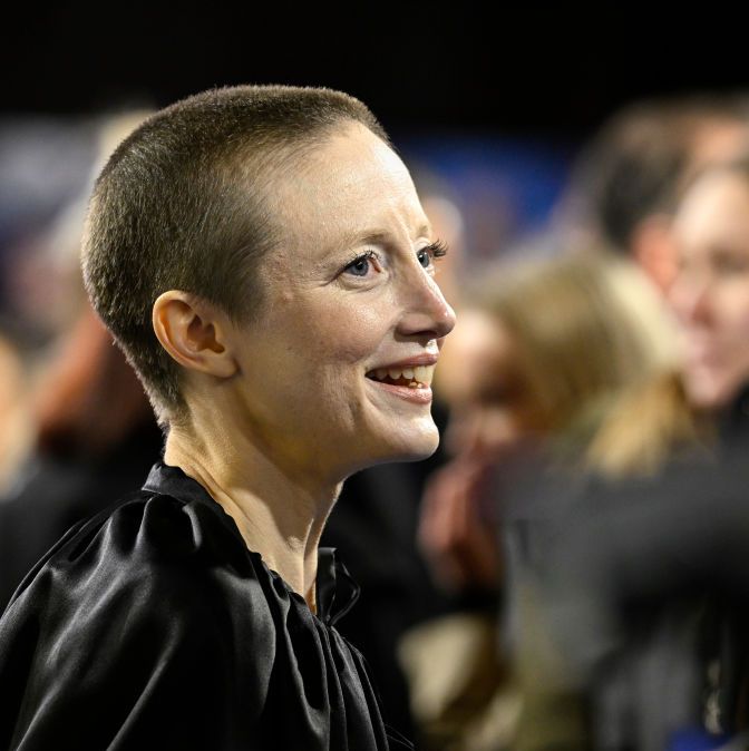 andrea riseborough, shown in profile looking toward the right of the frame, is smiling and wearing a black cape, the background is blurred out but shapes of people behind andrea are visible