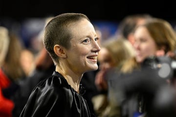 andrea riseborough, shown in profile looking toward the right of the frame, is smiling and wearing a black cape, the background is blurred out but shapes of people behind andrea are visible