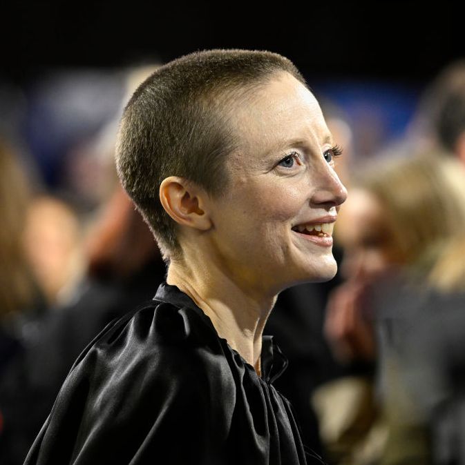 andrea riseborough, shown in profile looking toward the right of the frame, is smiling and wearing a black cape, the background is blurred out but shapes of people behind andrea are visible