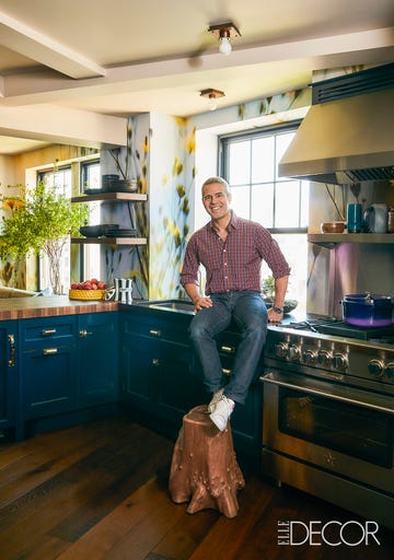 andy cohen sitting on a kitchen counter with legs resting on a side table made of a wood trunk