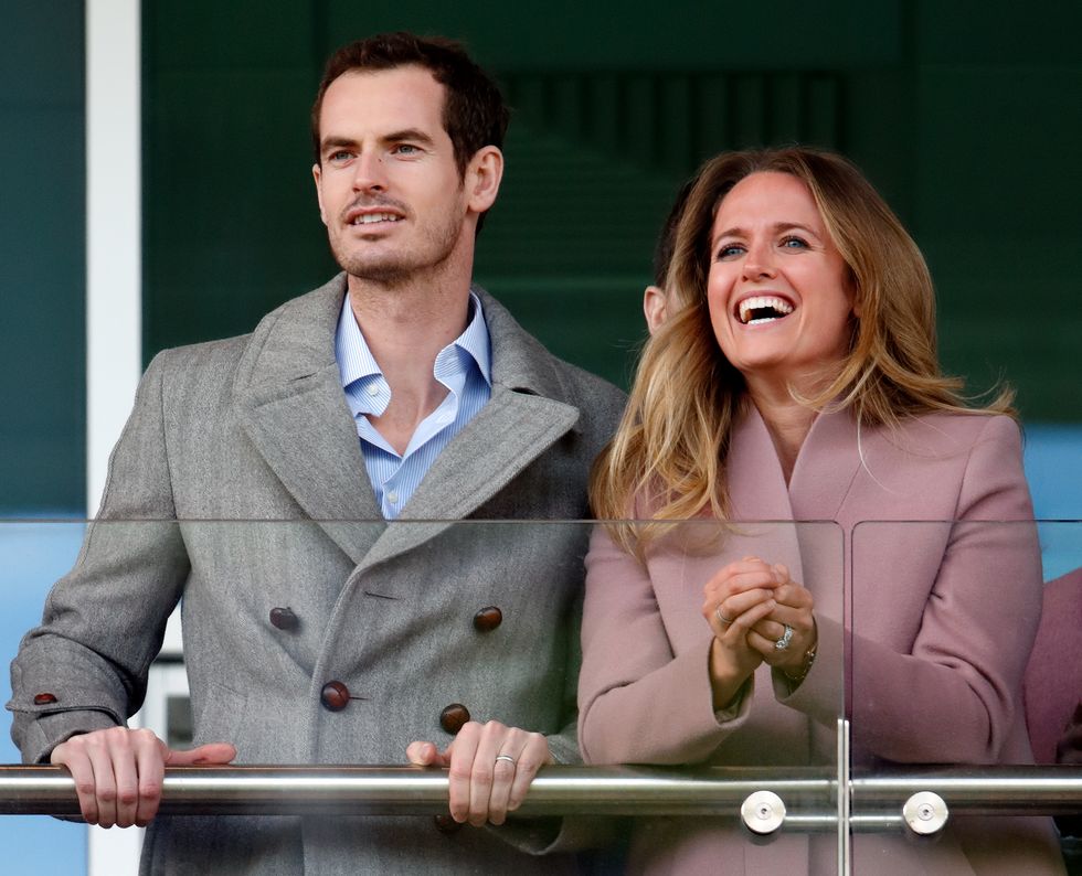 andy murray and kim sears stand behind a glass railing and look past the camera, both are wearing coats, he is holding onto the railing and she smiles and clasps her hands