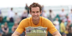 andy murray smiles at the camera while holding a silver bowl trophy, he wears an orange t shirt and leans against a tennis net