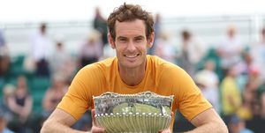andy murray smiles at the camera while holding a silver bowl trophy, he wears an orange t shirt and leans against a tennis net