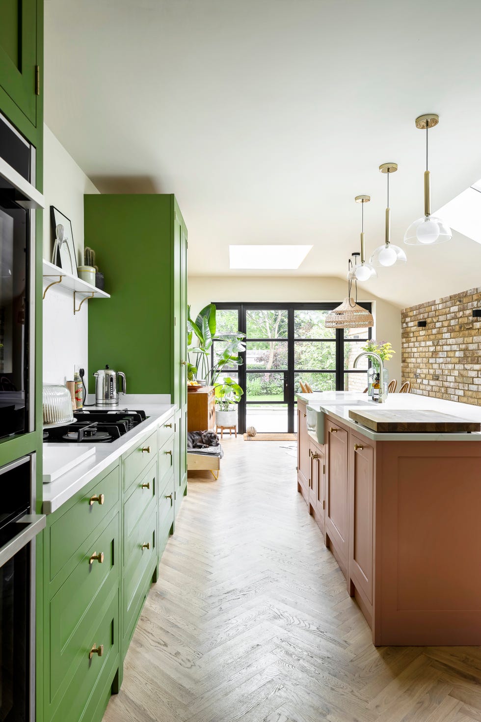 green kitchen units and pink island in room with black glazed doors overlooking a garden