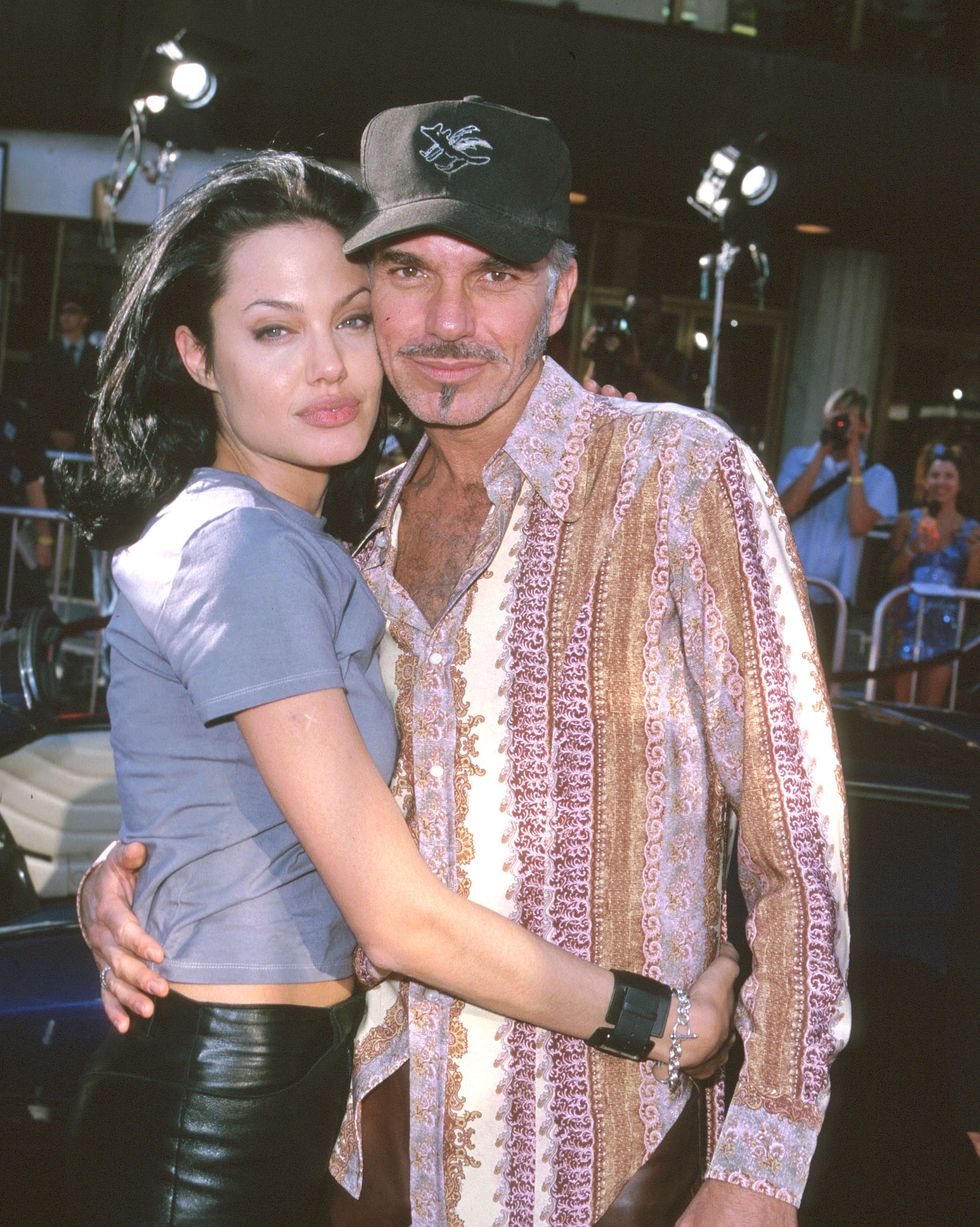 angelina jolie and billy bob thornton embrace while posing for photos, she wears a tshirt and black pants, he wears a black hat, purple brown and white paisley shirt and brown pants