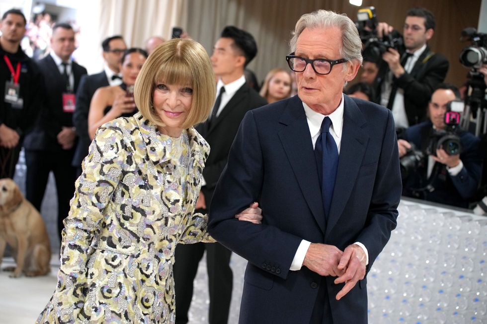 anna wintour, wearing a gray dress with black and yellow designs, walks arm in arm with bill nighy, wearing a black suit, blue tie, and glasses