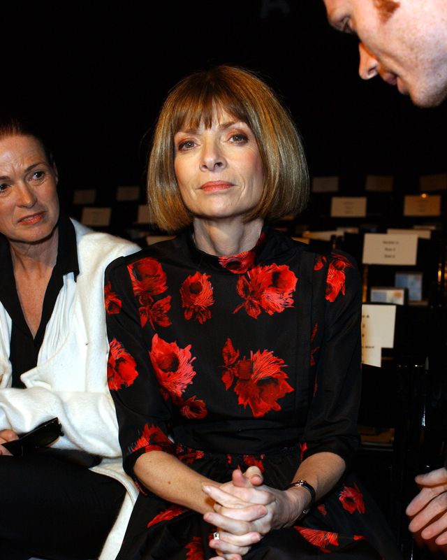 anna wintour looks at the camera with a neutral expression on her face, she is seated and clasps her hands in her lap, she is wearing a black dress with a pattern of large red flowers