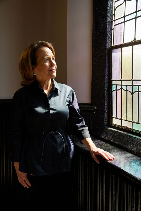 anne barrett doyle wearing a black blouse standing near a stained glass window with light shining on her face, highlighting her shoulder length auburn hair and green eyes