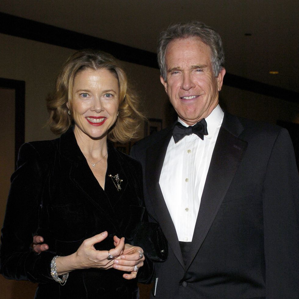 annette bening and warren beatty stand together and smile at the camera, she wears a black long sleeve dress with jewelry, he wears a black tuxedo with a white collared shirt