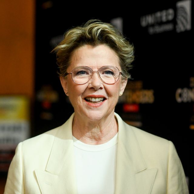 annette bening smiles toward the camera while standing in front of a dark background, she wears a yellow suit jacket, white shirt and aviator glasses