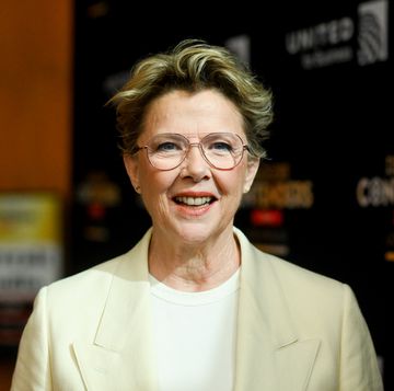 annette bening smiles toward the camera while standing in front of a dark background, she wears a yellow suit jacket, white shirt and aviator glasses