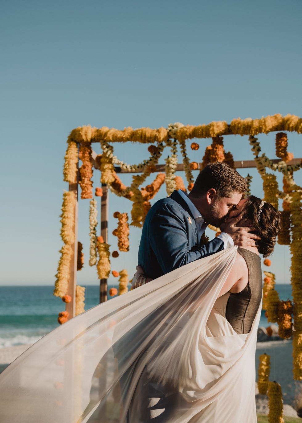 a bride in a custom black and white gown kisses a groom in a blue suit against a seaside background as her sheer white train whips in the wind