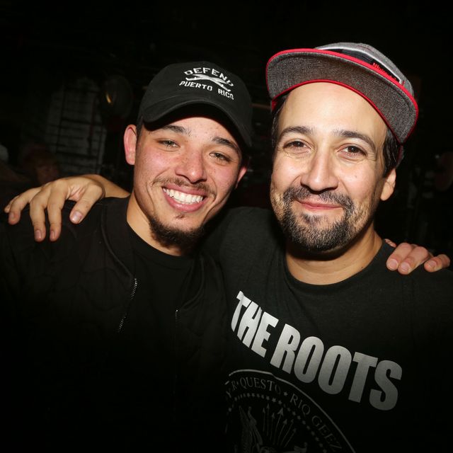 anthony ramos and lin manuel miranda, both wearing black t shirts and baseball caps, put their arms on each other's shoulders and smile for the camera