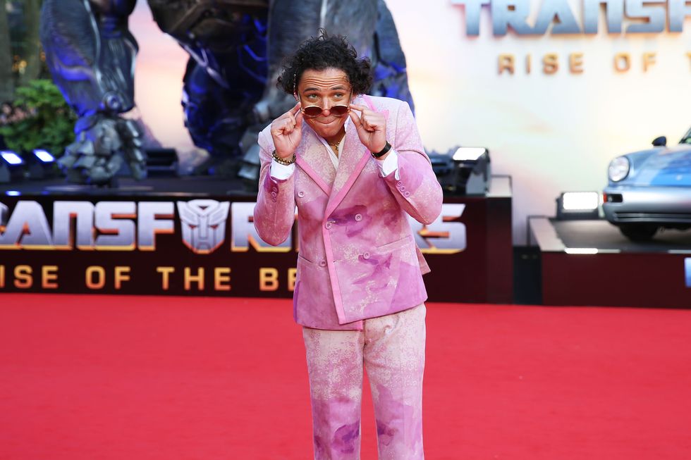 anthony ramos, wearing a pink suit and sunglasses, smiles and looks offscreen while lowering his sunglasses, standing on a red carpet in front of an advertisement for transformers rise of the beasts