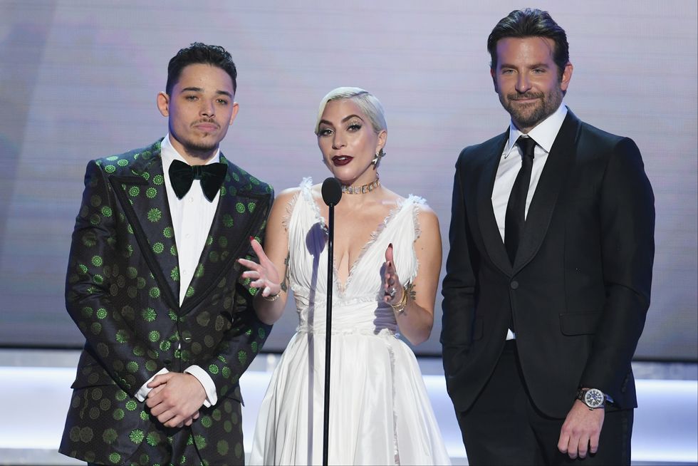 anthony ramos, wearing a black and green tuxedo, stands on a stage next to lady gaga, who wears a white dress and speaks into a microphone, and bradley cooper, wearing a black suit and tie